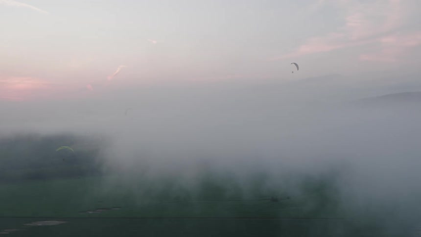 Aerial view of three paragliders soaring through a thick layer of fog blanketing a rural landscape at dawn