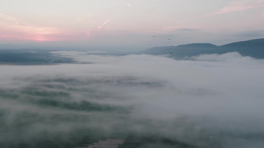 Paragliders soar peacefully above a dreamy, cloud-filled valley at sunrise, flying through the misty air