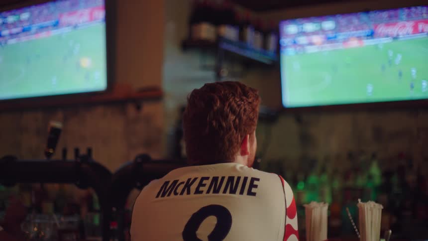 A focused man in a soccer jersey sits at a bar, intently watching a game on two television screens