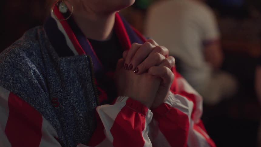 A woman in a colorful retro jacket clasps her hands together in suspense while watching an event