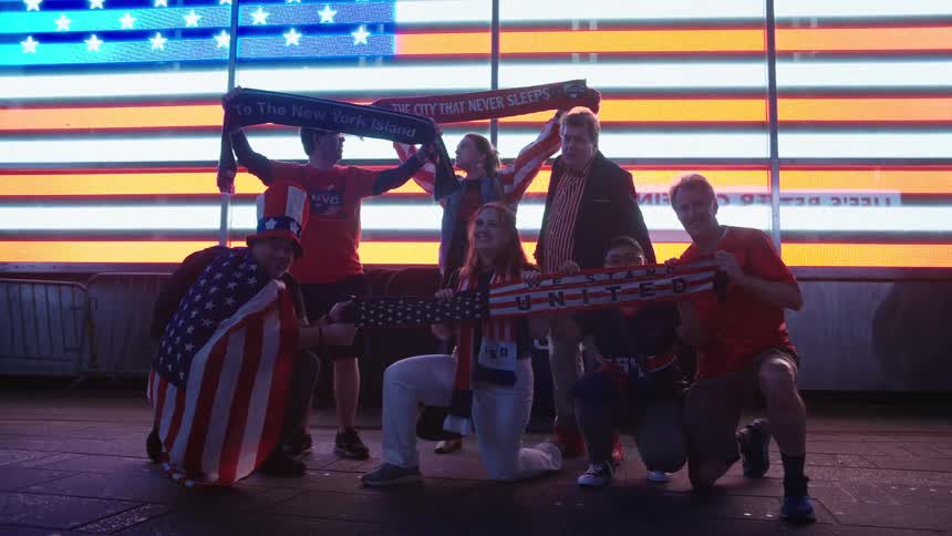 Patriotic fans dressed in red, white, and blue celebrate and pose with scarves in front of a giant American flag display in Times Square