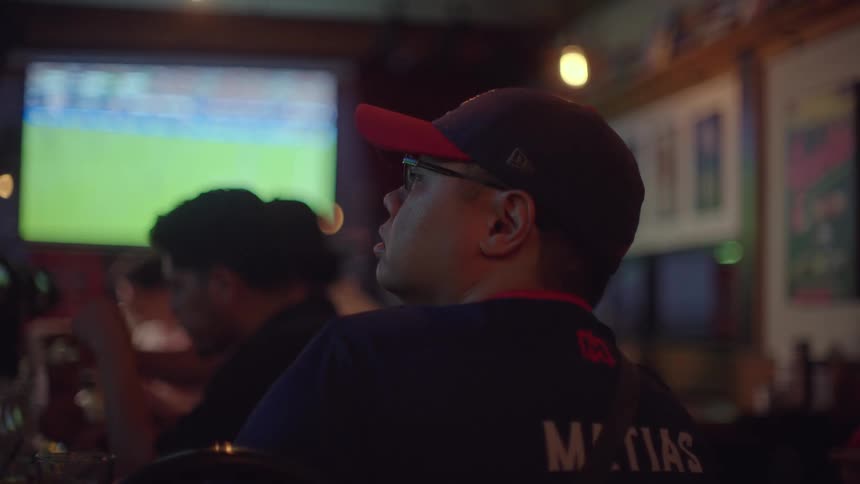 A man wearing a baseball cap intently watches a soccer game on a big screen in a dimly lit sports bar