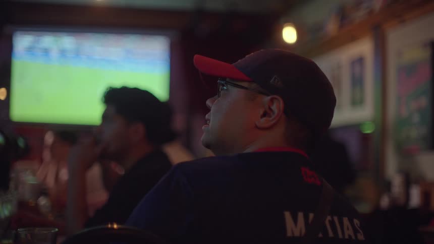 A man wearing a baseball cap intently watches a sports game on a projector screen in a crowded bar
