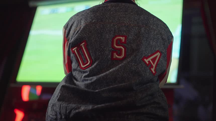 A patriotic sports fan wearing a USA jacket watches a soccer game on television from behind