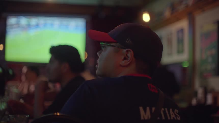 A young man wearing a baseball cap and glasses intently watches a sports game on a large screen inside a dark, crowded bar