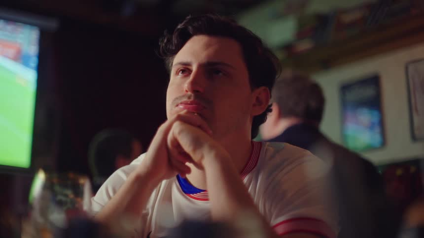 An anxious soccer fan with his hands clasped sits in a bar watching a game on TV