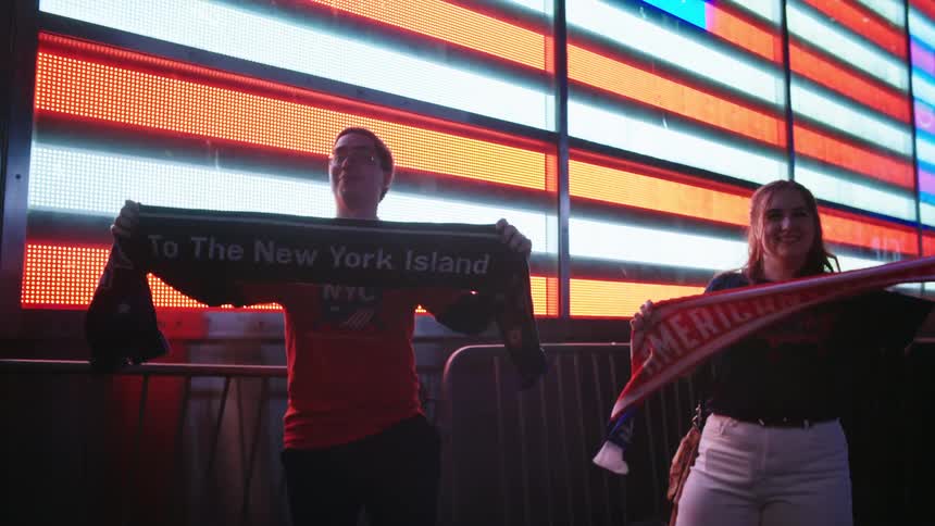 Two patriotic American sports fans proudly display their team scarves in slow motion in front of a massive illuminated American flag in Times Square