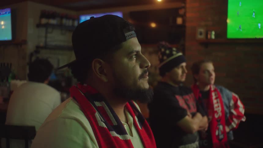 A focused man with a beard drinks a beer in a sports bar while intensely watching a soccer match on television with fellow fans