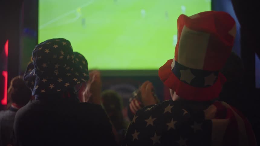 From behind, a crowd of American sports fans wearing patriotic hats cheer and clap while watching a soccer game on TV