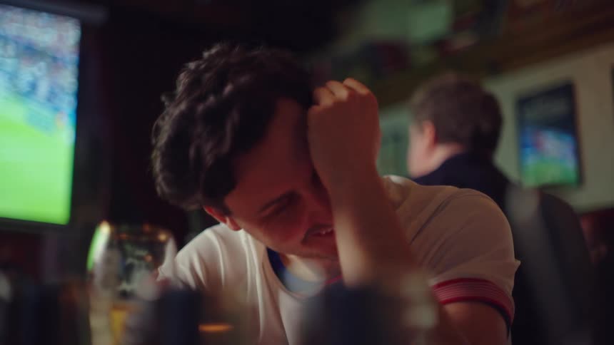 A male sports fan in a bar experiences a rollercoaster of emotions, from despair to laughter, while watching a game on TV