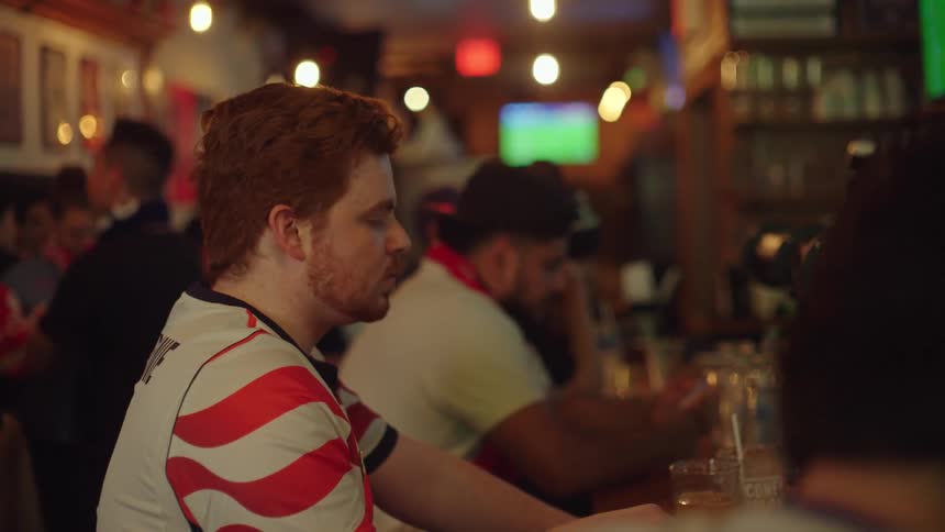 A group of male sports fans intently watch a soccer game at a bar, with one man reacting with excitement in slow motion