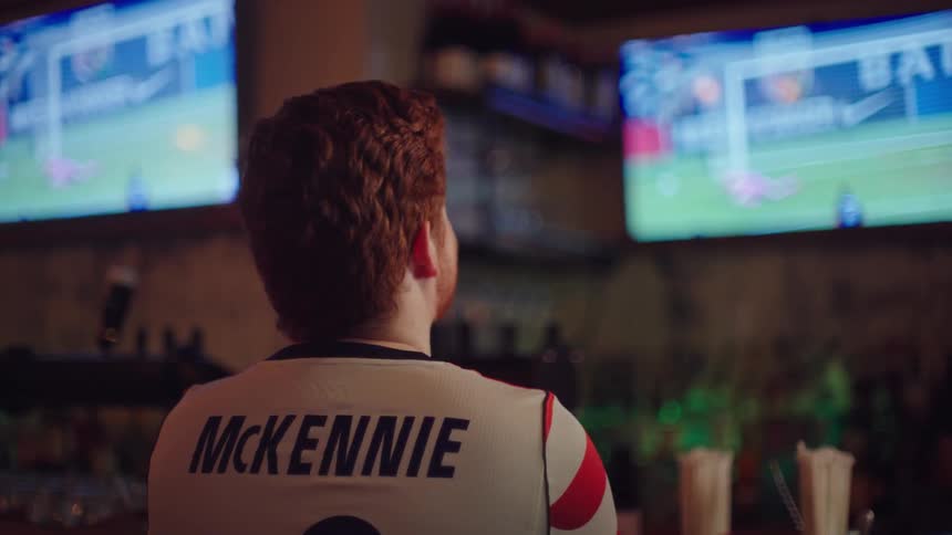 A man with red hair wearing a soccer jersey sits at a bar intently watching a game on two television screens