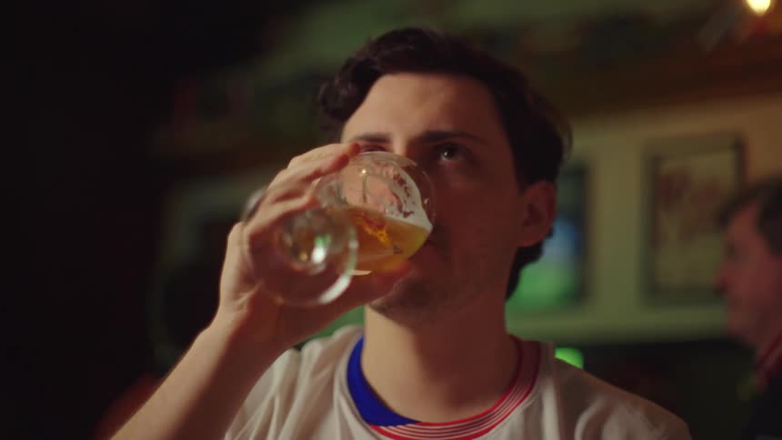 A tense soccer fan in a USA jersey anxiously watches the game while drinking a beer in a sports bar