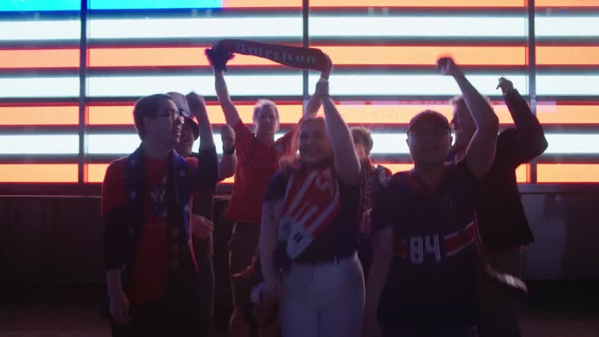 A diverse group of American fans wildly celebrate a victory, jumping and cheering in front of a massive digital American flag display in the city at night