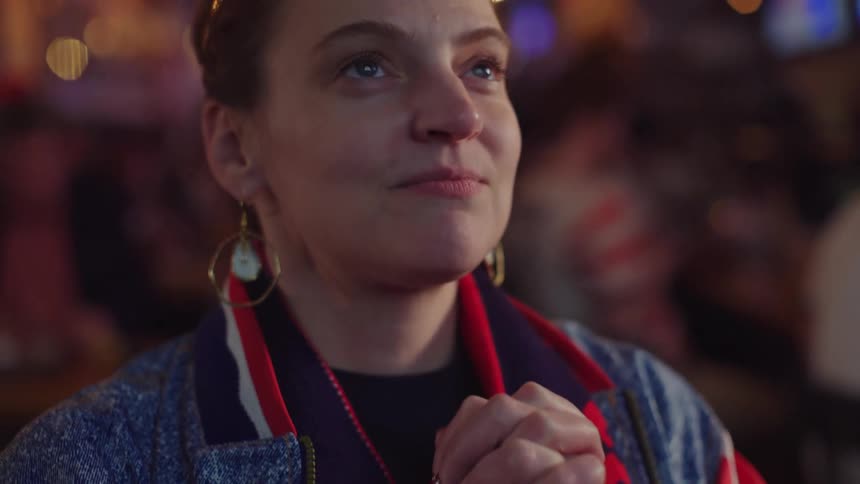 A woman in a retro jacket clasps her hands together in hopeful anticipation while watching a sports game at a bar
