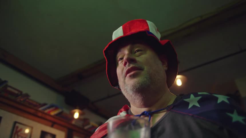 A patriotic American superfan dressed in an Uncle Sam hat and a flag cape reacts with disappointment while watching a game