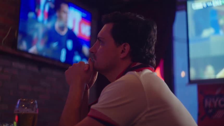 A lone male sports fan with a beer watches a game with intense focus and anxiety at a sports bar