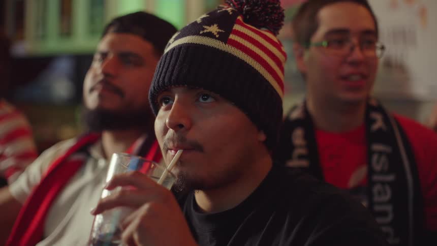 A patriotic young man intently watches a televised sports match while drinking with friends at a bar
