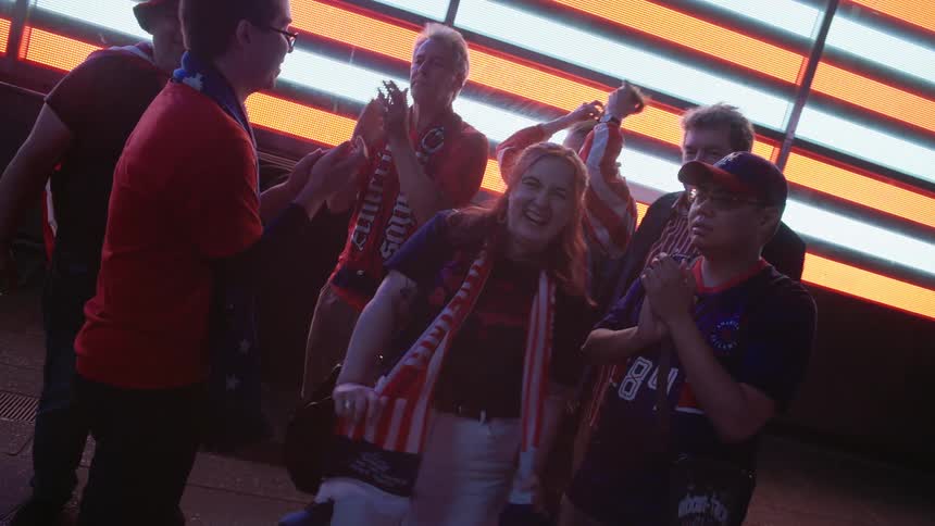 A diverse group of patriotic American fans cheer and celebrate together in slow motion at night in front of a giant illuminated flag