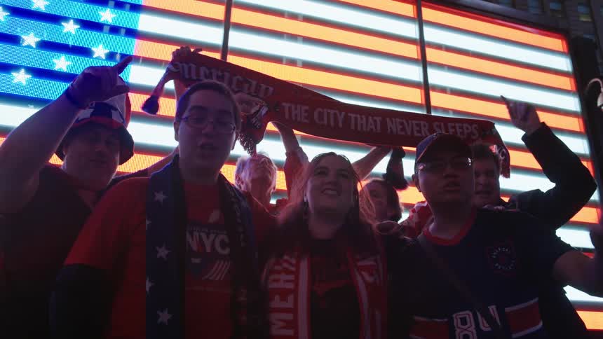 A diverse group of patriotic sports fans cheer and sing together in Times Square in front of a giant illuminated American flag