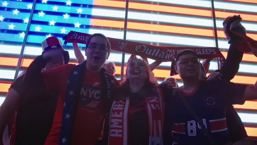 A group of patriotic sports fans celebrate together at night in Times Square, cheering in front of a giant illuminated American flag billboard