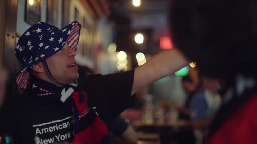 A slow-motion close-up of a man in a patriotic American flag hat and scarf talking with a friend in a bar