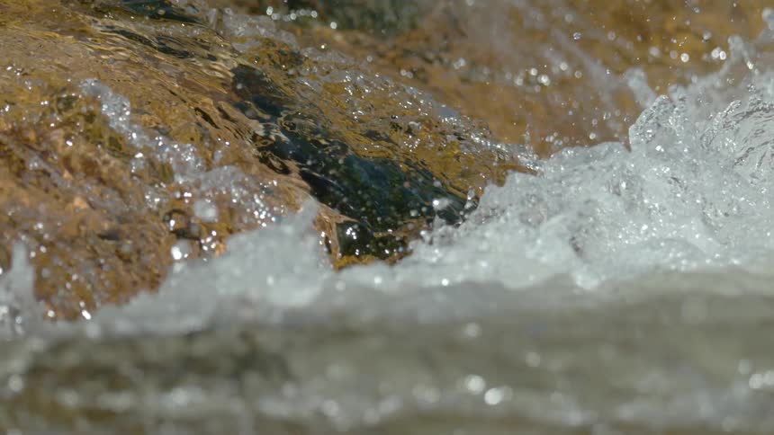 A super slow-motion close-up of a powerful stream of water cascading over sunlit golden rocks