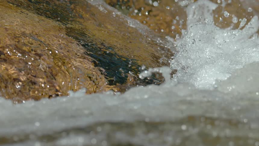 Crystal clear water cascades over smooth, golden rocks in a sunlit river, captured in mesmerizing super slow motion