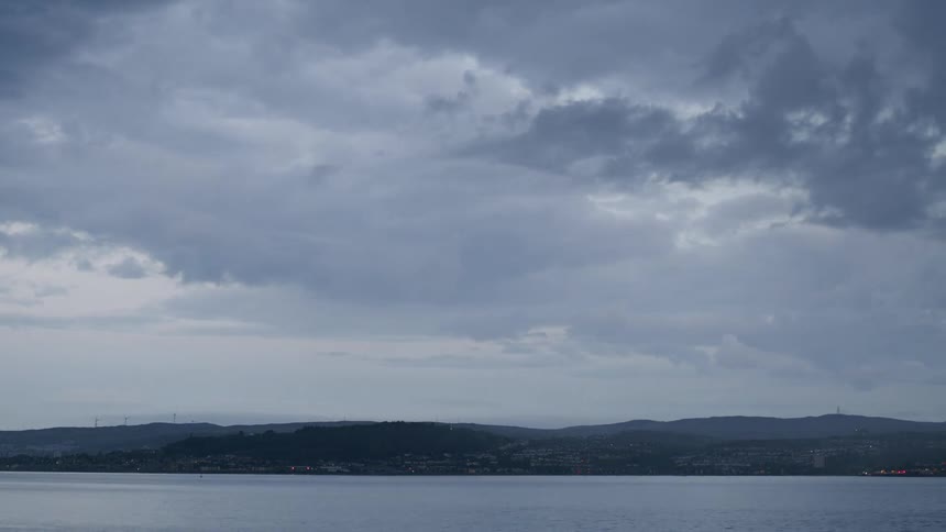 A dramatic time-lapse of ominous grey clouds rolling over a tranquil body of water and a distant coastal town as evening falls