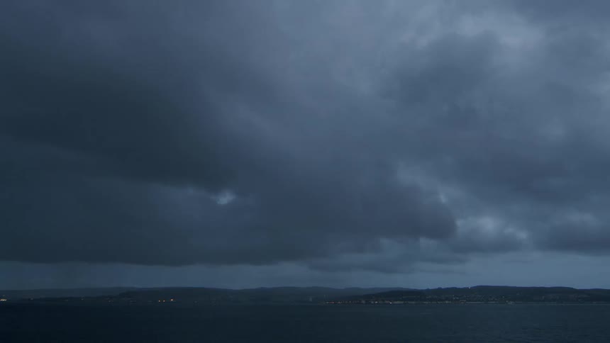 A dramatic time-lapse shows dark storm clouds sweeping across the ocean sky above a distant coastal city at dusk with heavy rain