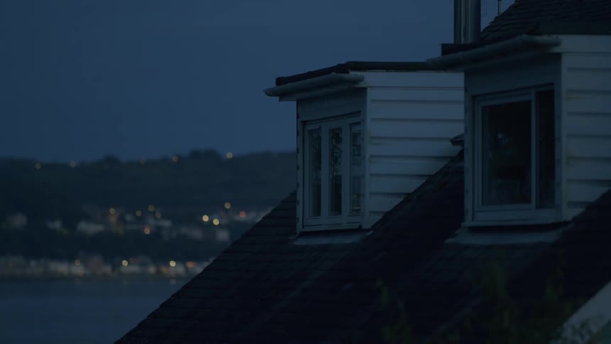 Dormer windows on a rooftop at dusk, with the blurred lights of a coastal town across the water in the background
