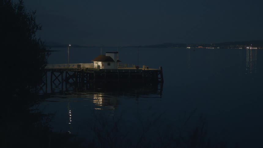 A tranquil pier rests on a calm bay at night as distant city lights reflect on the water's surface