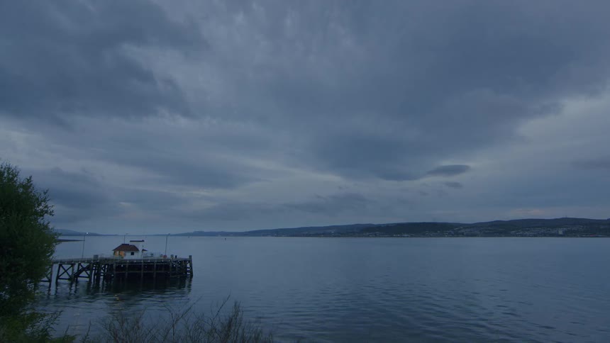 A dramatic timelapse shows dark, ominous clouds passing over a tranquil bay with a small pier as day fades into night