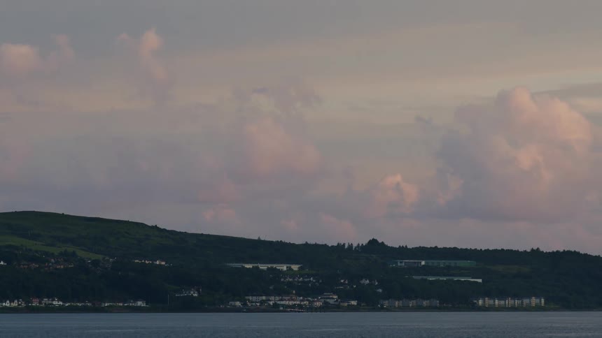 A tranquil time-lapse shows soft, pink-hued clouds drifting over a seaside town nestled along a green coastline
