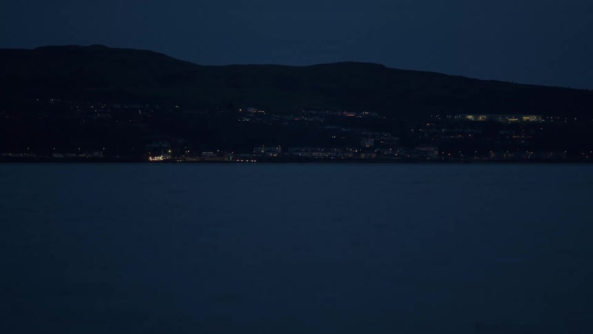 A tranquil wide shot of a coastal town illuminated by lights at night across a calm body of water
