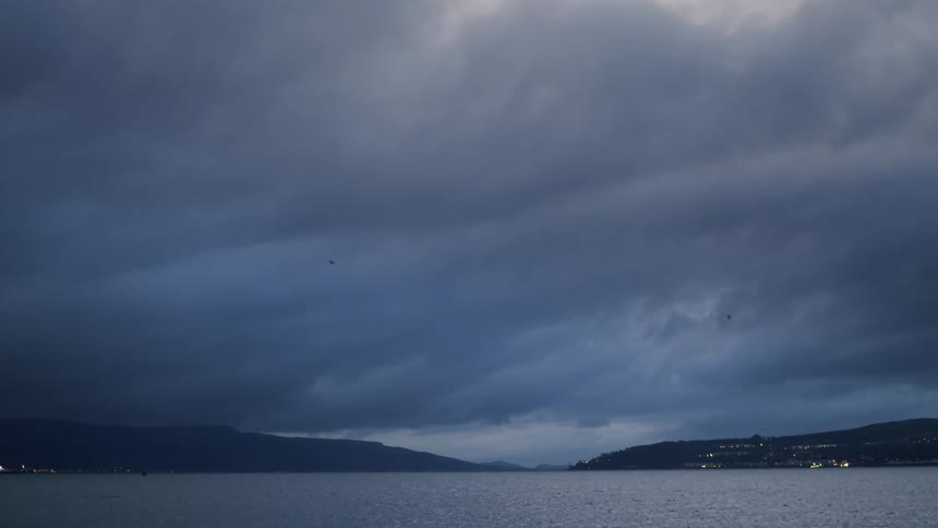 Dark, moody storm clouds roll over a calm body of water at twilight as birds fly across the ominous sky