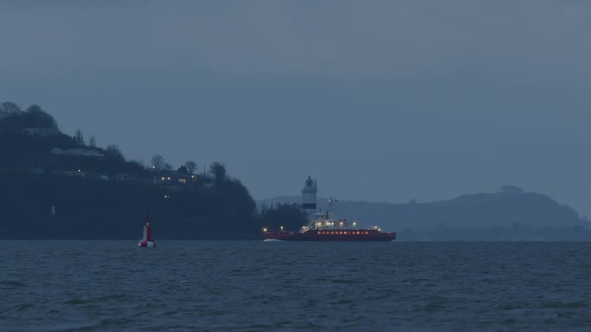 A red ferry slowly sails past a coastal lighthouse and a navigation buoy during a gloomy dusk