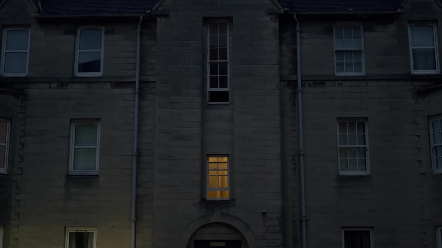 A static shot of a large, old stone building at dusk, where only a single window glows with a warm light, creating a mysterious atmosphere