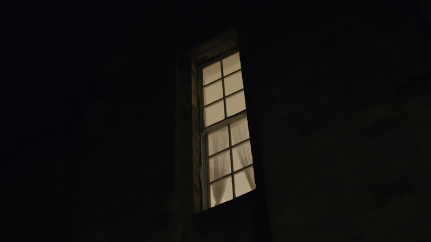 A low-angle shot captures a warm, glowing window on a dark building facade at night
