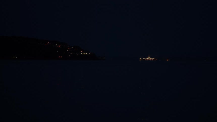 An illuminated ship floats on the calm dark water at night with the lights of a distant shore visible in the background