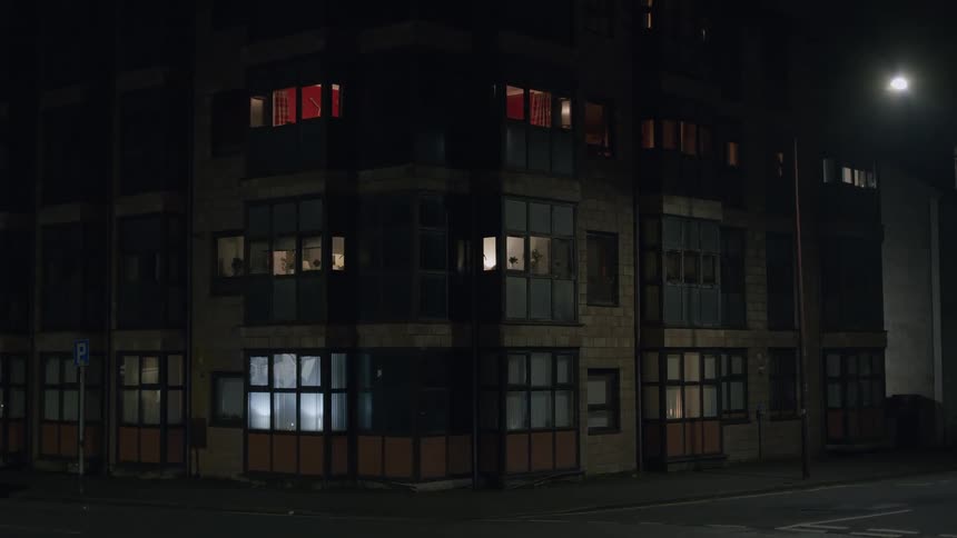 The glowing windows of a city apartment building illuminate a quiet street corner at night as a car passes