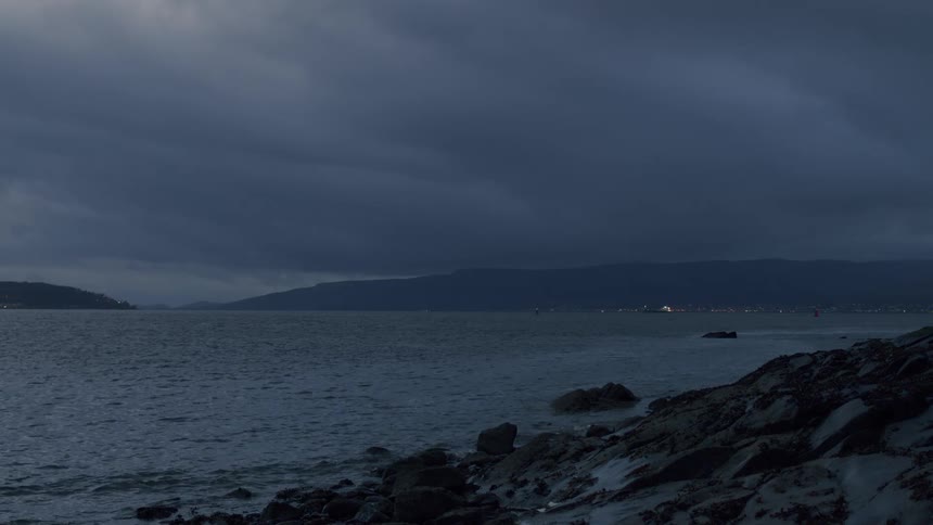 Stormy clouds gather over a dark sea at twilight, with the lights of a distant city glowing on the horizon
