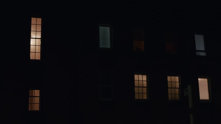 Windows of a city apartment building glow with warm light against the dark facade at night
