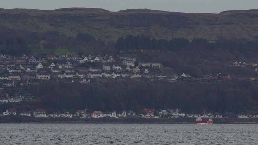 A white car ferry sails past a small coastal town nestled at the base of a green hill on a quiet, overcast day