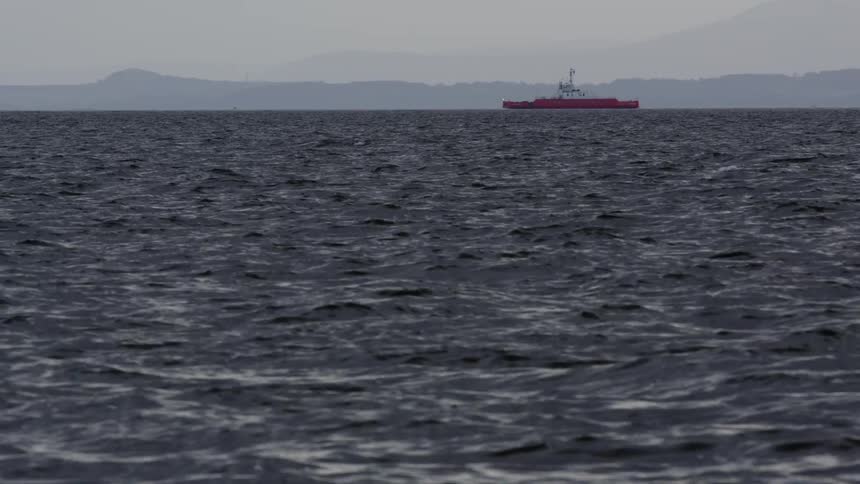 A red ferry ship passes in the distance on a dark, choppy sea with a hazy mountain range on the horizon