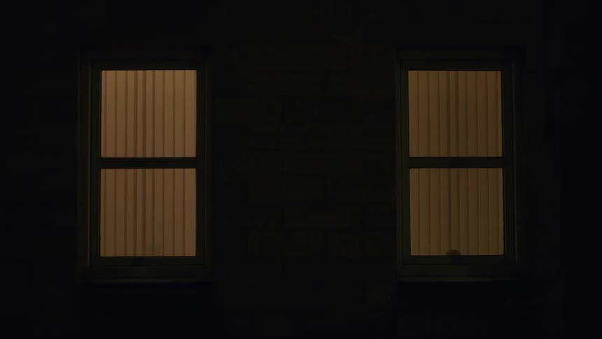 Mysterious lights from passing cars reflect in two warmly lit windows on a dark night