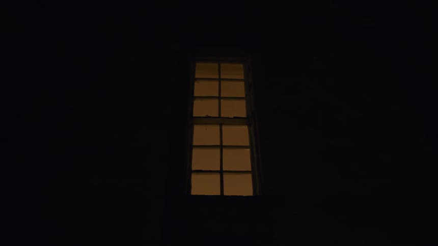 A handheld shot shows a single, tall window glowing with warm light surrounded by a completely dark building at night