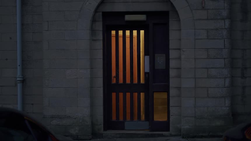 Warm light glows from behind a dark wooden door of a stone building at night