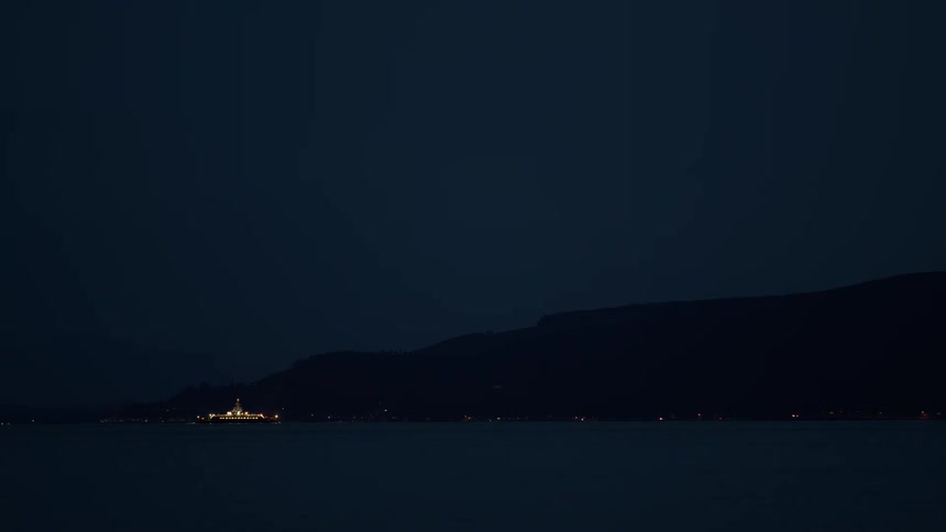 A brightly lit ferry boat glides across the dark water at night in front of a silhouetted shoreline