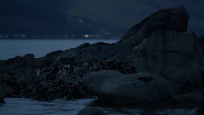 A static shot of a rocky shoreline at dusk as calm waters lap against the stones with a distant hillside in the background